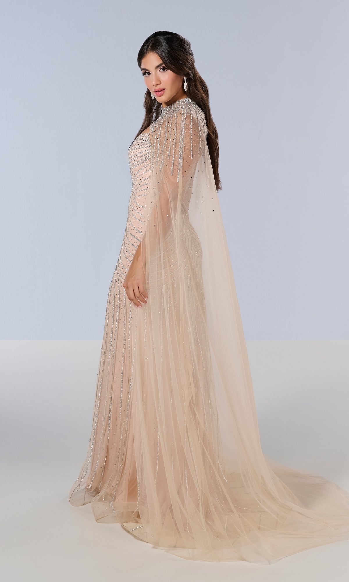 A woman wears the Tiffany 16202 long prom dress—a floor-length, embellished tulle gown with sparkling details and a sheer cape—against a plain light background.
